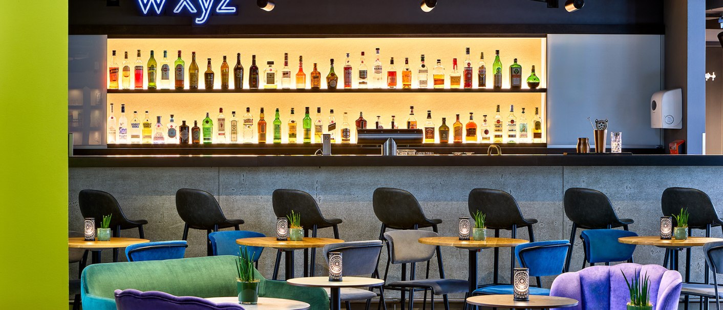 Moderne Bar mit bunten Sesseln und beleuchtetem Flaschenregal. Der Schriftzug "WXYZ" leuchtet &uuml;ber der Theke., &copy; Aloft Stuttgart