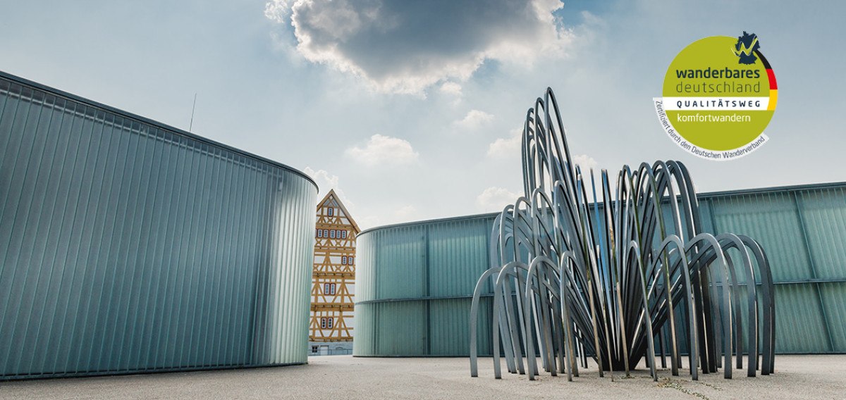 Moderne Skulptur vor einem Fachwerkhaus in Waiblingen. Im Hintergrund Wolken am Himmel. Logo von 'Wanderbares Deutschland' oben rechts., © Unbekannt