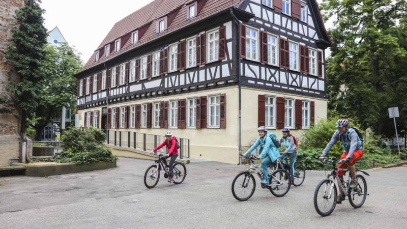 Vier Radfahrer fahren an einem historischen Fachwerkhaus in Kirchheim unter Teck vorbei. Das Gebäude hat weiße Wände und braune Fensterläden., © Schwäbische Alb Tourismusverband e.V. Vier Radfahrer fahren an einem historischen Fachwerkhaus in Kirchheim unter Teck vorbei. Das Gebäude hat weiße Wände und braune Fensterläden., © Schwäbische Alb Tourismusverband e.V.