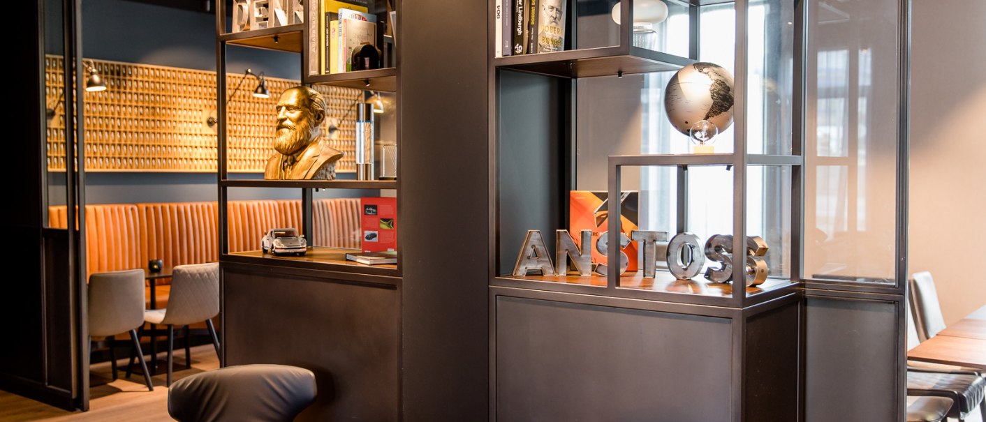 Modernes Café mit Regalen, Büchern, einer Büste und einem Globus. Sitzbereich mit braunen Polstern und stilvollen Stühlen., © TOMAS Modernes Café mit Regalen, Büchern, einer Büste und einem Globus. Sitzbereich mit braunen Polstern und stilvollen Stühlen., © TOMAS
