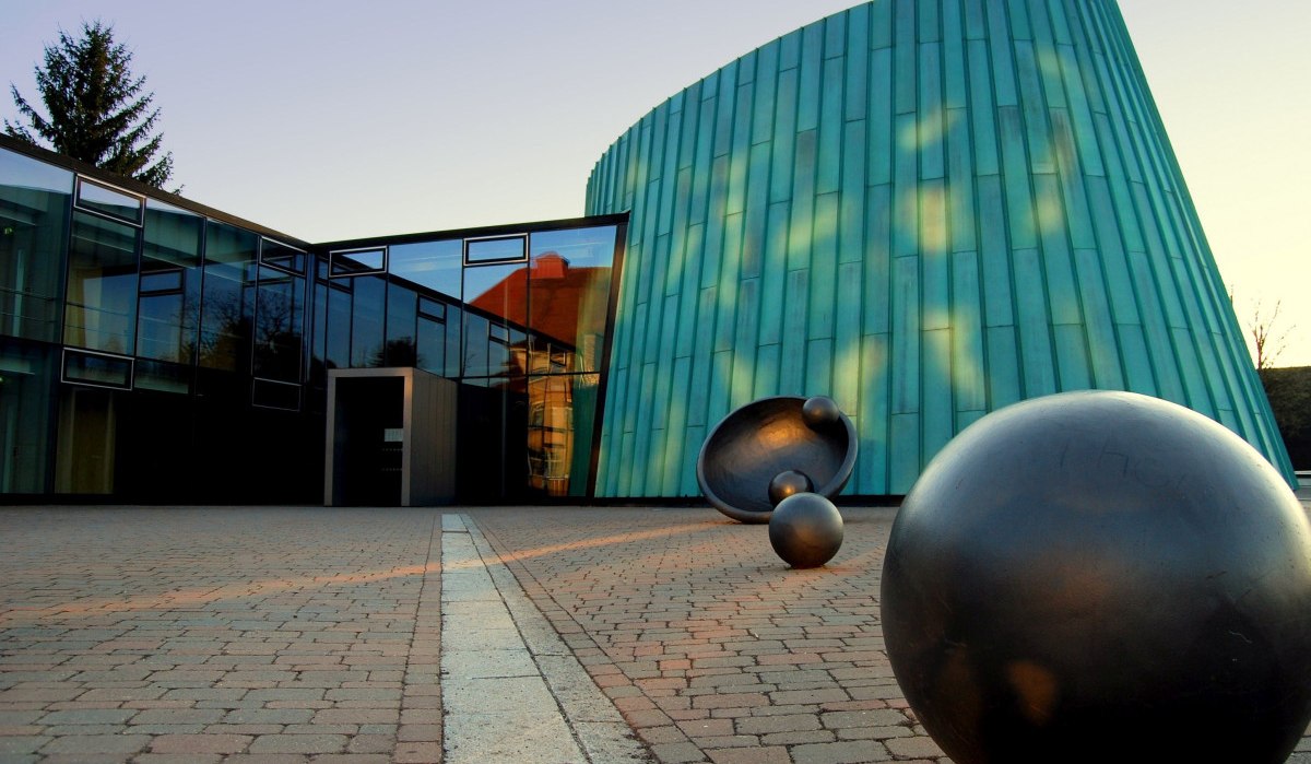 Moderne Architektur der Musikschule Fellbach mit Glasfassade und grüner Metallverkleidung. Im Vordergrund sind schwarze Kugelskulpturen zu sehen., © Simone Mathias Moderne Architektur der Musikschule Fellbach mit Glasfassade und grüner Metallverkleidung. Im Vordergrund sind schwarze Kugelskulpturen zu sehen., © Simone Mathias