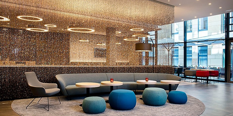 Moderne Lobby mit grauem Sofa, blauen Hockern und runden Tischen vor einer glitzernden Wand., © Holiday Inn Express Waiblingen