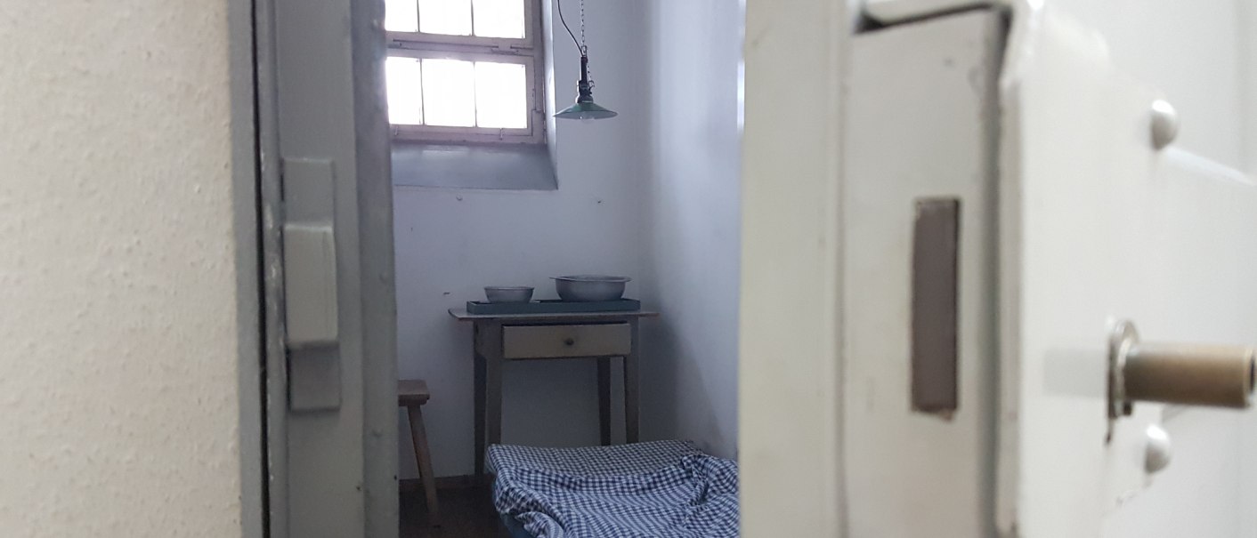 Blick in eine historische Gefängniszelle mit einfachem Bett, kleinem Tisch und Schüsseln. Ein Fenster mit Gitterstäben lässt Licht herein., © Strafvollzugsmuseum Ludwigsburg