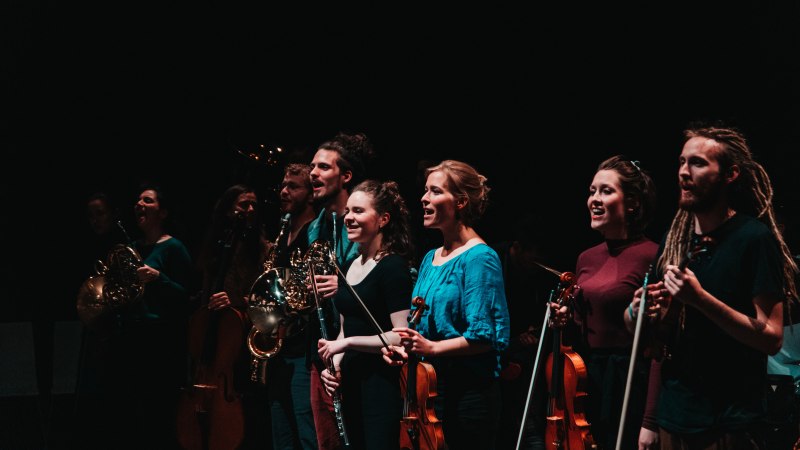 Musiker auf einer B&uuml;hne mit H&ouml;rnern, Geigen und Fl&ouml;ten, in dunkler Umgebung, l&auml;chelnd und konzentriert., &copy; Sky B&uuml;rhaus