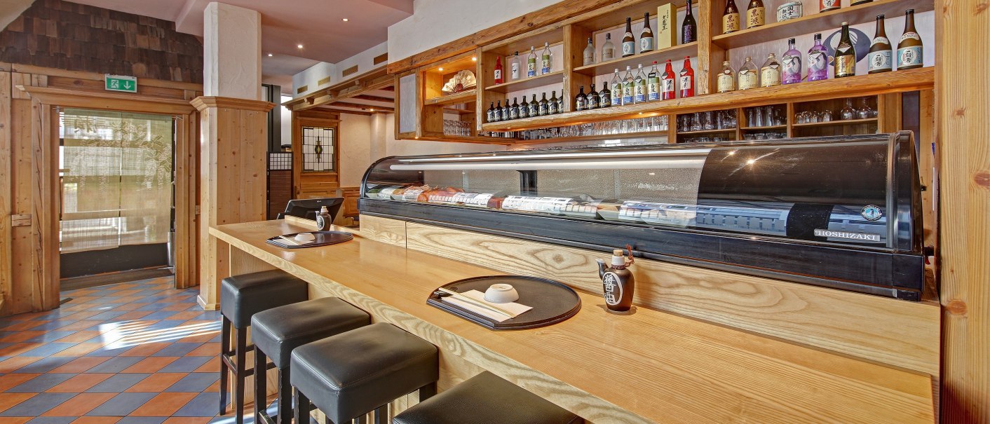 Ein japanisches Restaurant mit Sushi-Theke, Barhockern und Regalen voller Sake-Flaschen. Holzelemente dominieren das Interieur., © Restaurant Kurose
