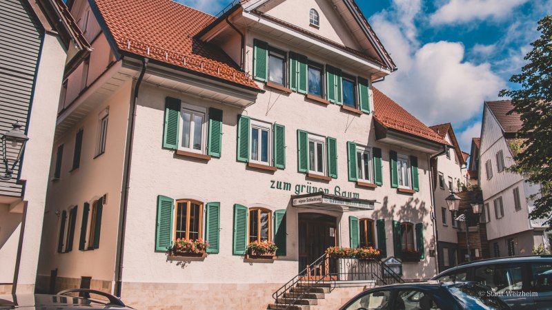 Ein traditionelles Gasthaus mit grünen Fensterläden und roten Dachziegeln, umgeben von anderen Gebäuden. Der Himmel ist blau mit einigen Wolken., © Stadt Welzheim Ein traditionelles Gasthaus mit grünen Fensterläden und roten Dachziegeln, umgeben von anderen Gebäuden. Der Himmel ist blau mit einigen Wolken., © Stadt Welzheim