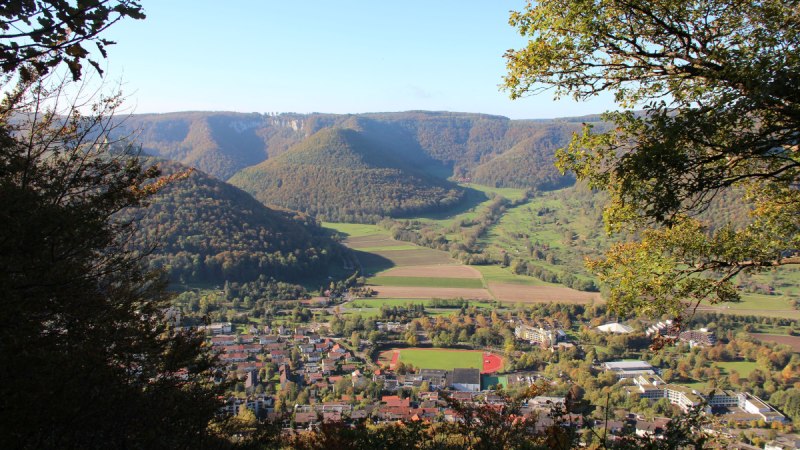 Ausblick vom Nägelesfelsen auf eine Stadt mit Sportplatz, umgeben von grünen Hügeln und Wäldern unter klarem Himmel., © Bad Urach Tourismus