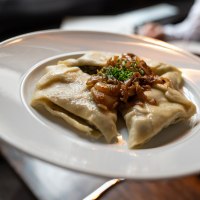 Ein Teller mit Maultaschen, garniert mit geschmolzenen Zwiebeln und Schnittlauch, wird serviert. Im Hintergrund ist eine Person unscharf zu sehen., © Stuttgart-Marketing GmbH, Martina Denker