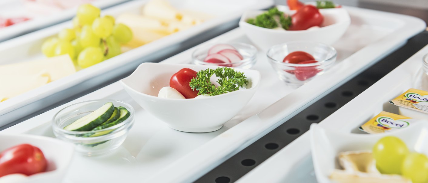 Ein Frühstücksbuffet mit Schalen voller Tomaten, Gurken, Radieschen, Trauben und Butterportionen auf weißen Tabletts., © TOMAS