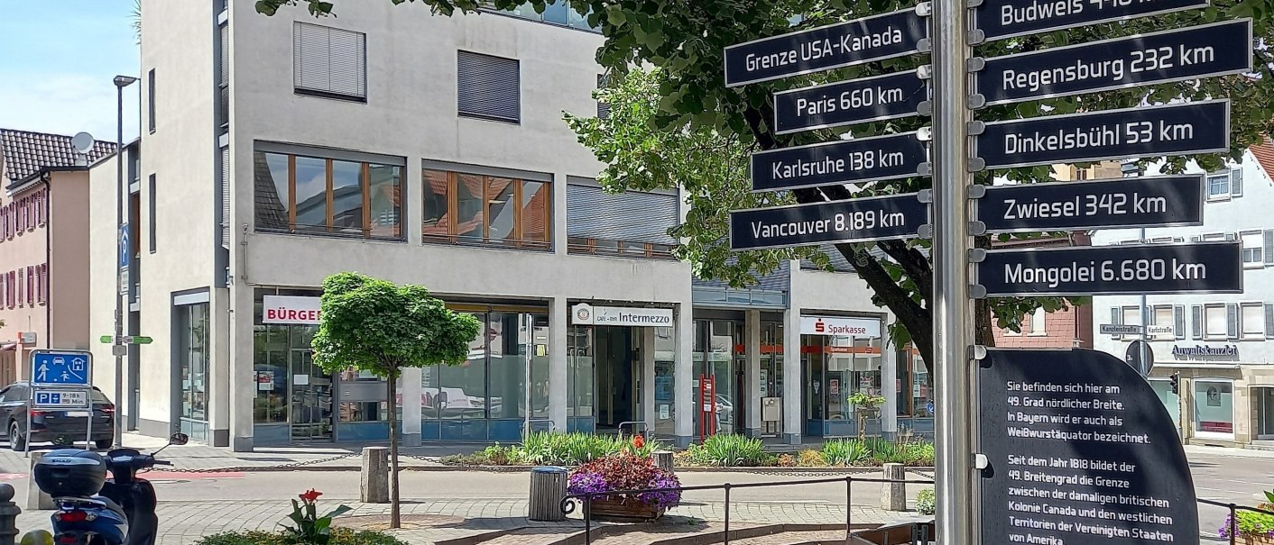 Wegweiser auf einem Platz mit Entfernungen zu Städten und Orten auf dem 49. Breitengrad, wie Paris und Vancouver. Im Hintergrund ein modernes, weißes Gebäude, © Petra Natzkowski Wegweiser auf einem Platz mit Entfernungen zu Städten und Orten auf dem 49. Breitengrad, wie Paris und Vancouver. Im Hintergrund ein modernes, weißes Gebäude, © Petra Natzkowski