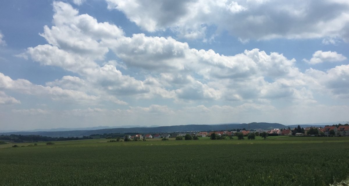 Weite Felder erstrecken sich vor einem Dorf, das am Horizont liegt. Der Himmel ist mit Wolken bedeckt, die Landschaft wirkt friedlich und weitl&auml;ufig., &copy; www.pro-cycl.de