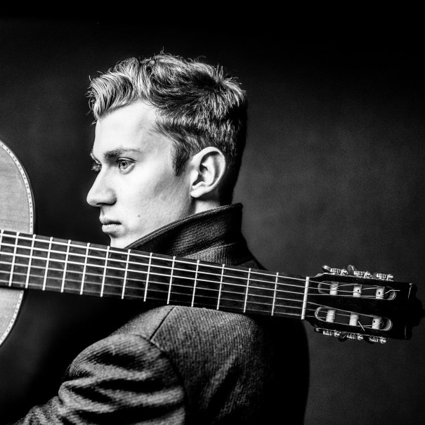 Schwarz-weiß Porträt eines Mannes mit Gitarre über der Schulter, im Profil. Er trägt einen hochgeschlossenen Mantel., © Marco Borggreve