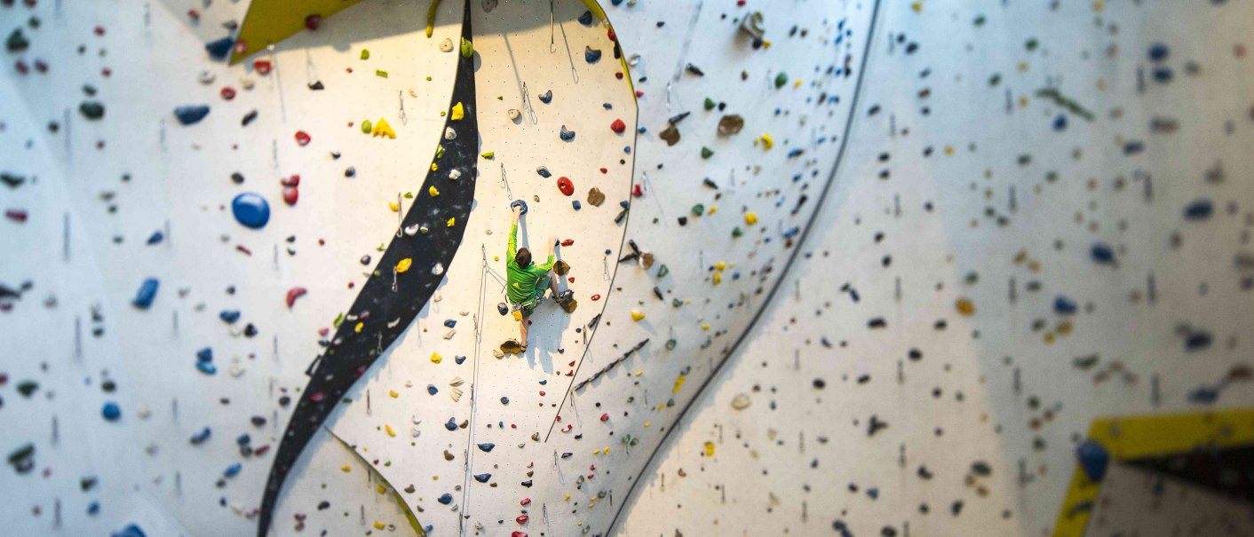 Ein Kletterer in grüner Kleidung erklimmt eine hohe Indoor-Kletterwand mit bunten Griffen im DAV Kletter- und Boulderzentrum Schwaben., © Rockerei