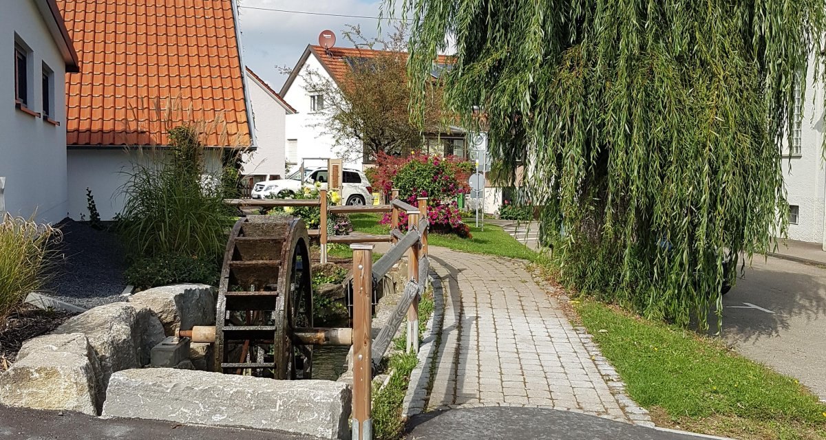 Ein Mühlrad an einem Wasserkanal neben einem gepflasterten Weg, umgeben von Häusern und einem großen Baum in Mögglingen., © Remstal Tourismus e.V. Ein Mühlrad an einem Wasserkanal neben einem gepflasterten Weg, umgeben von Häusern und einem großen Baum in Mögglingen., © Remstal Tourismus e.V.
