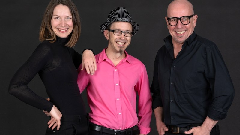 Drei Personen in schwarzer und pinker Kleidung posieren lächelnd vor einem dunklen Hintergrund. Eine Frau und zwei Männer stehen nebeneinander., © Tanz- und Theaterwerkstatt e.V.