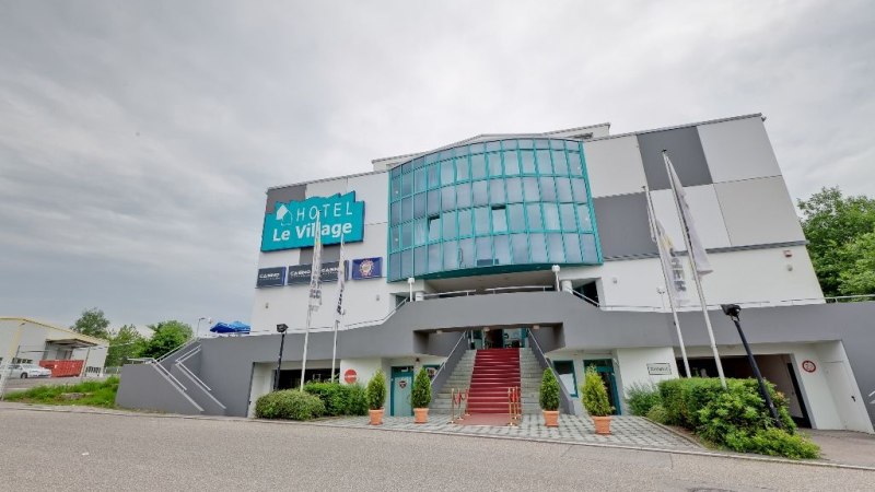 Das Hotel Le Village zeigt eine moderne Fassade mit Glasfront und Treppe. Vor dem Gebäude wehen Fahnen, der Himmel ist bewölkt., © Hotel Le Village Das Hotel Le Village zeigt eine moderne Fassade mit Glasfront und Treppe. Vor dem Gebäude wehen Fahnen, der Himmel ist bewölkt., © Hotel Le Village