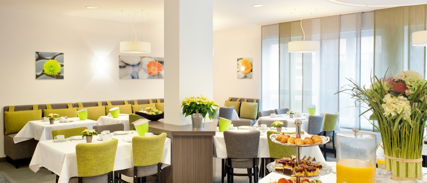 Heller Frühstücksraum mit gedeckten Tischen, grünen Stühlen und einem Buffet mit Saft, Gebäck und Obst. Blumen und Bilder schmücken den Raum., © TOMAS