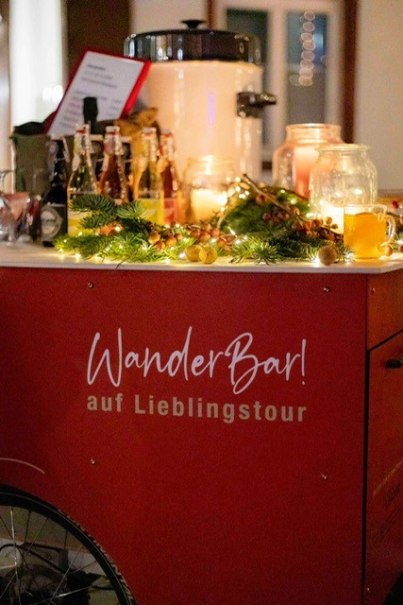 Ein mobiler Getr&auml;nkestand namens 'WanderBar!' ist festlich mit Lichtern und Tannenzweigen dekoriert. Verschiedene Getr&auml;nke sind auf dem Stand platziert.