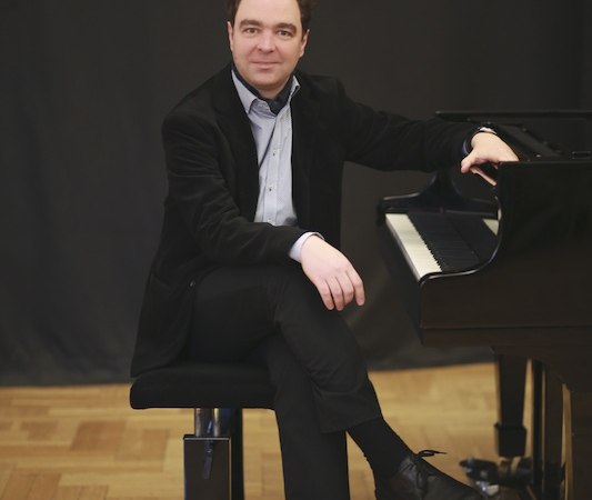 Ein Mann in Anzug sitzt lächelnd auf einem Klavierhocker neben einem Flügel, vor einem dunklen Hintergrund., © Stadt Böblingen - Amt für Kultur