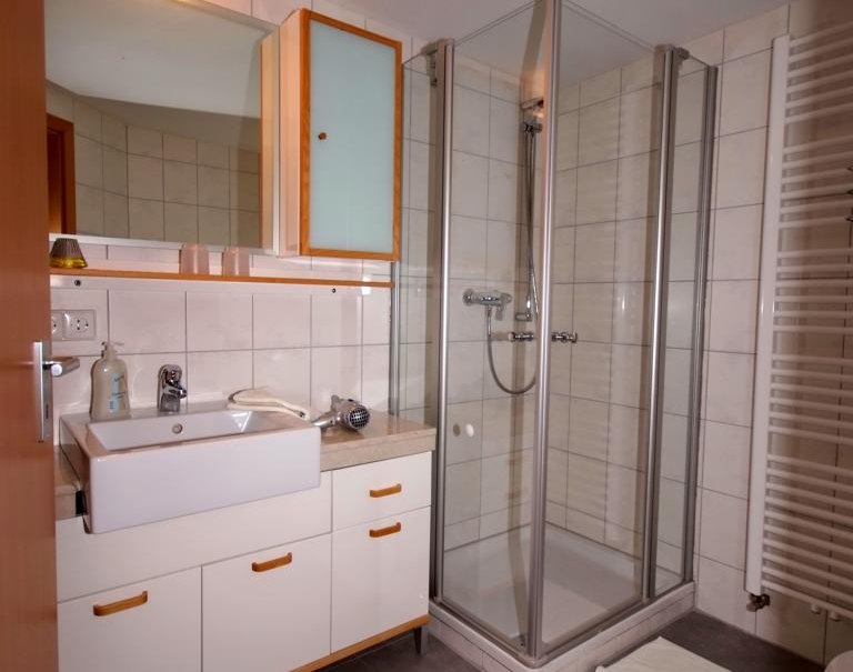 Modernes Badezimmer mit Dusche, Waschbecken und Spiegel. Weiße Fliesen, grauer Boden, Handtuchheizkörper und Holzdetails prägen den Raum., © TOMAS