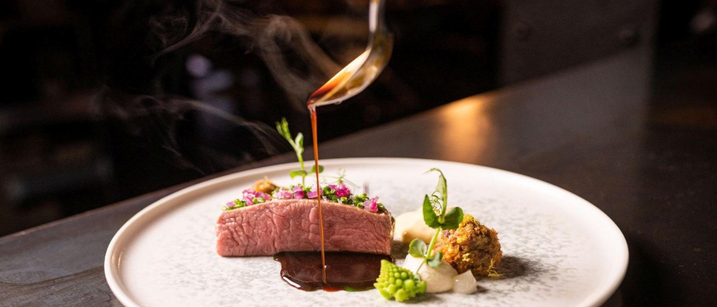 Ein kunstvoll angerichtetes Gourmetgericht mit Fleisch, Gemüse und einer dunklen Sauce, die aus einer Kelle auf den Teller gegossen wird., © 5 - Stuttgart