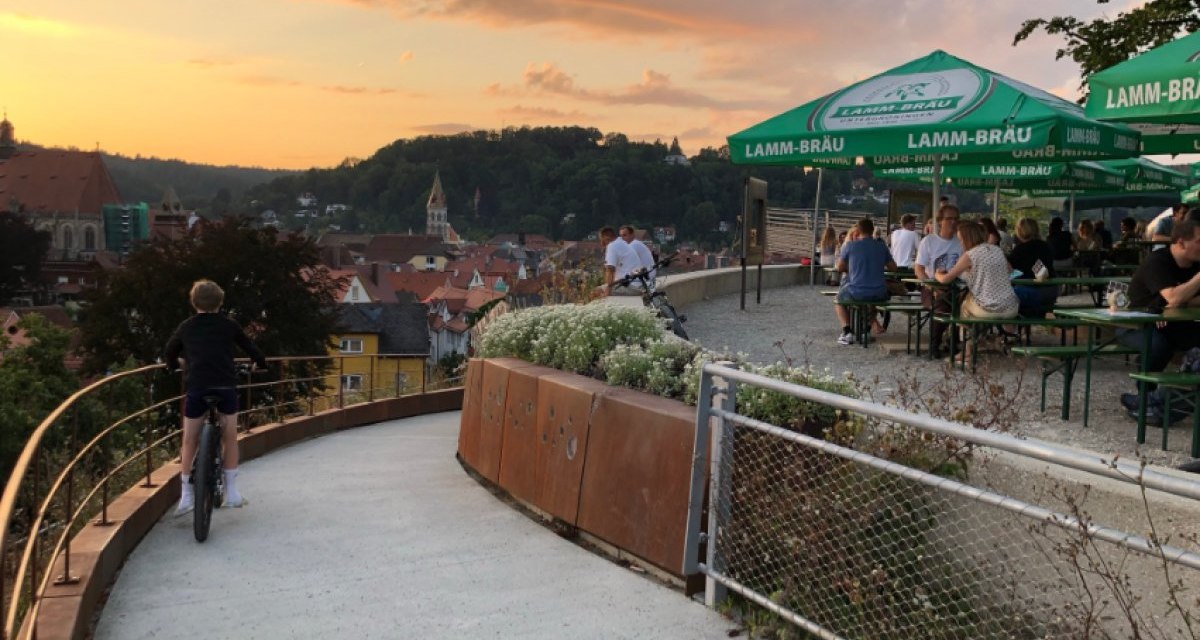 Biergarten mit grünen Sonnenschirmen und Gästen, ein Kind fährt Fahrrad auf einem Weg. Im Hintergrund eine Stadtansicht bei Sonnenuntergang., © Touristik und Marketing GmbH Biergarten mit grünen Sonnenschirmen und Gästen, ein Kind fährt Fahrrad auf einem Weg. Im Hintergrund eine Stadtansicht bei Sonnenuntergang., © Touristik und Marketing GmbH