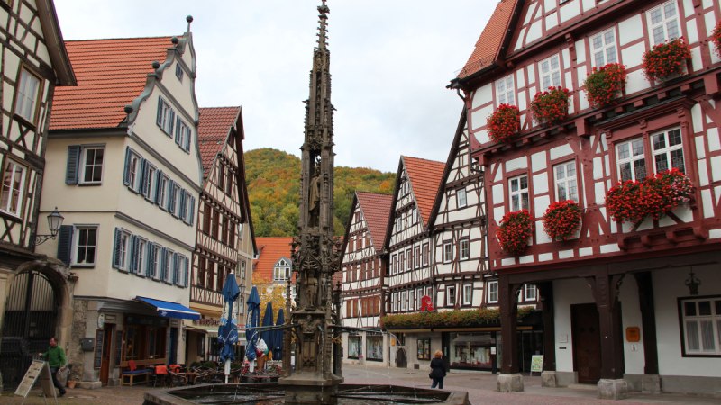 Ein malerischer Marktplatz mit Fachwerkh&auml;usern, einem verzierten Brunnen und herbstlich gef&auml;rbten B&auml;umen im Hintergrund.