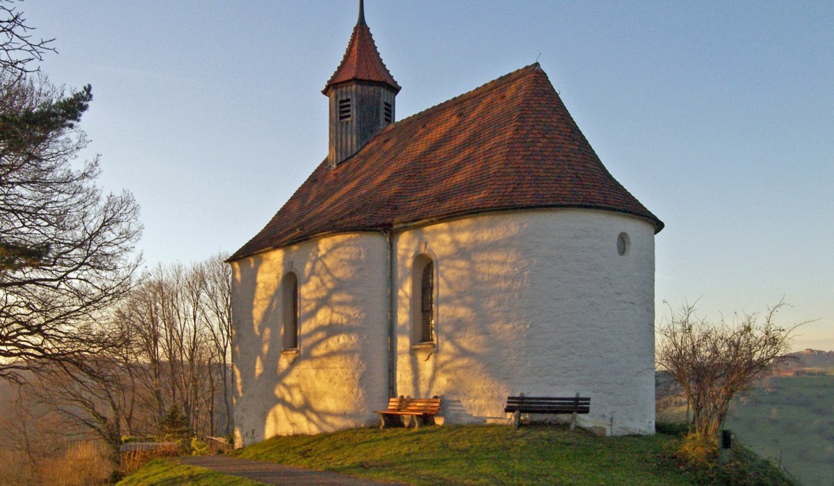 Die Marienkapelle Wißgoldingen im Abendlicht, umgeben von Bäumen. Schatten der Äste zeichnen sich auf der weißen Fassade ab. Zwei Bänke stehen davor., © Foto: Frieder Kopper