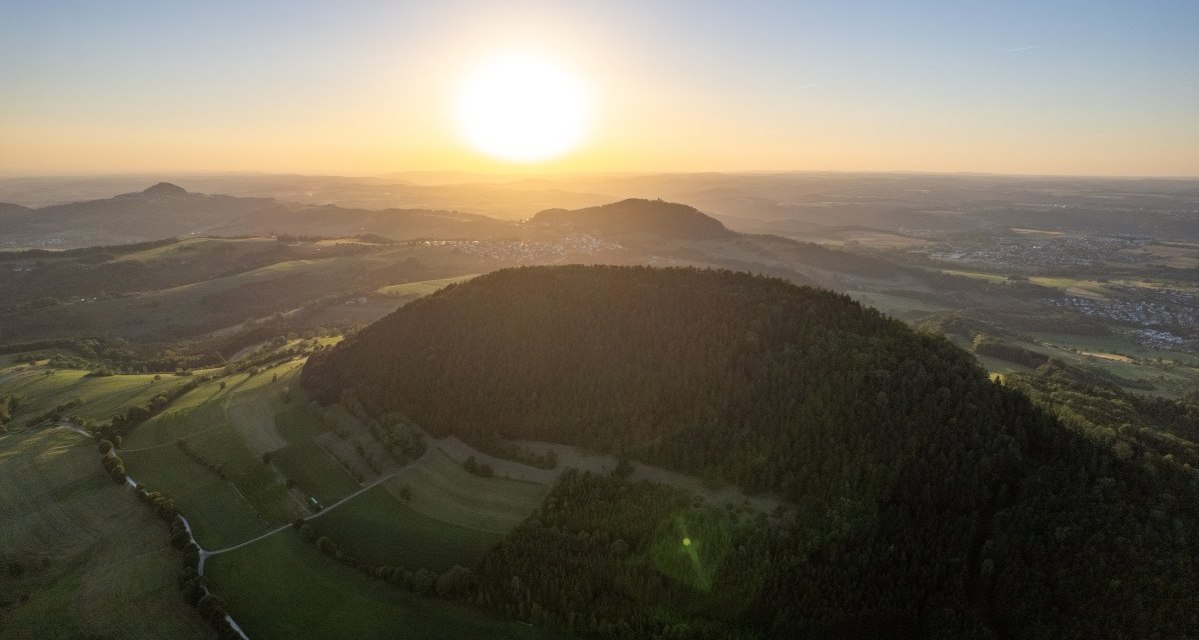 Luftaufnahme eines bewaldeten Hügels bei Sonnenuntergang, umgeben von Feldern und Dörfern in einer weiten Landschaft., © Foto Thomas Zehnder Luftaufnahme eines bewaldeten Hügels bei Sonnenuntergang, umgeben von Feldern und Dörfern in einer weiten Landschaft., © Foto Thomas Zehnder