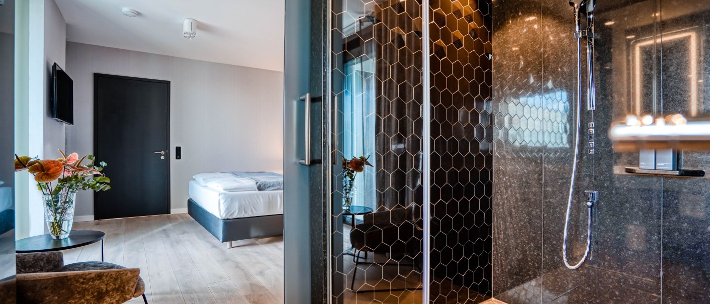 Moderne Hotelsuite mit elegantem Design, Bett, Glasdusche mit Wabenmuster, Blumen und stilvollen Möbeln auf Holzboden., © CLOUD N°7 APARTMENTS GmbH