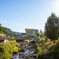 Moderne Architektur im Remspark, umgeben von Natur. Ein Fluss mit Brücke im Vordergrund, Bäume und ein Gebäude links., © Mario Klaiber