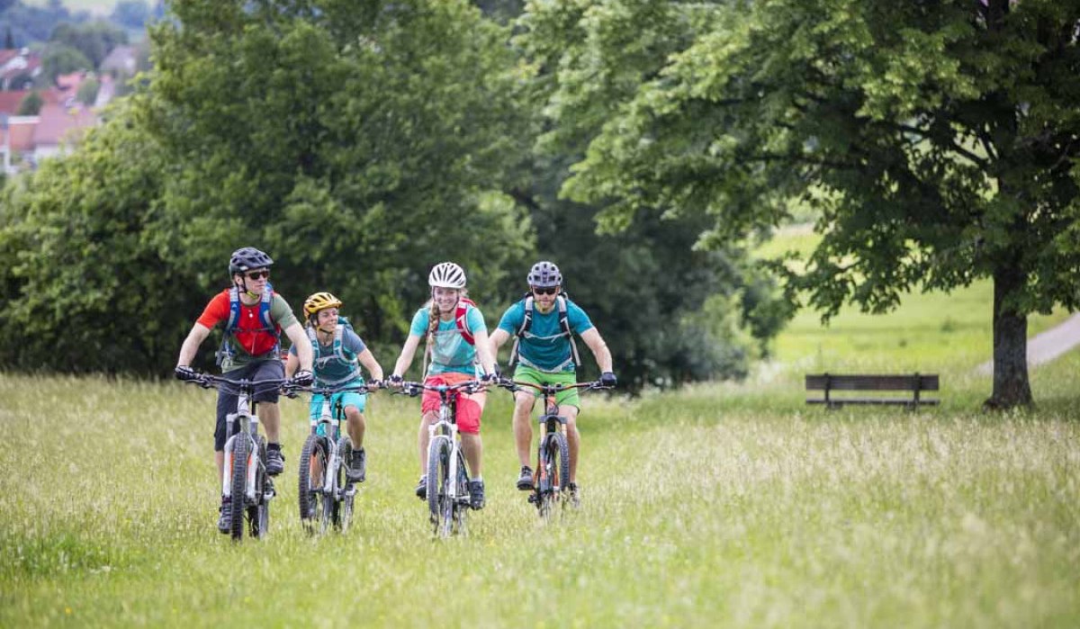 Vier Radfahrer fahren auf einem grasbewachsenen Weg, umgeben von Bäumen und Wiesen. Im Hintergrund sind Häuser und Hügel zu sehen., © Schwäbische Alb Tourismusverband e.V.