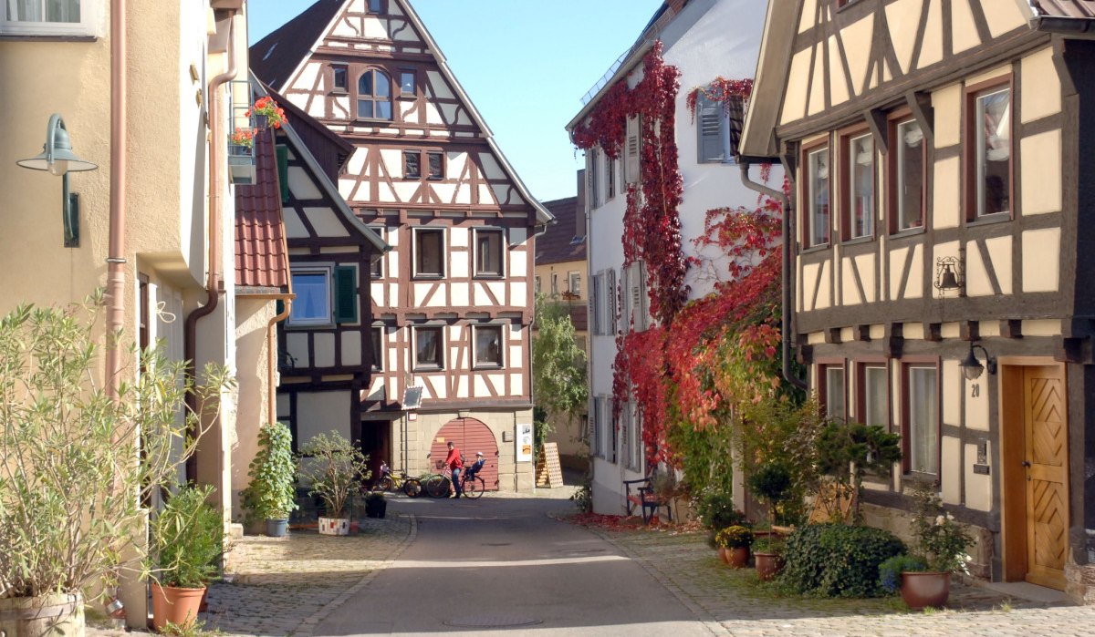Enge Gasse mit Fachwerkh&auml;usern, roten Weinreben an den W&auml;nden und Pflanzen in T&ouml;pfen. Zwei Personen mit Fahrr&auml;dern im Hintergrund., &copy; Stadt Herrenberg