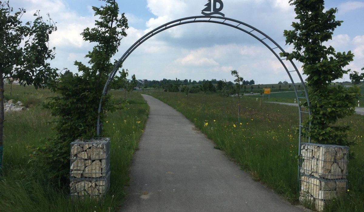 Ein Torbogen aus Metall mit einem Symbol oben markiert den Eingang zu einem Pfad. Der Weg führt durch eine grüne Wiese mit Bäumen., © www.pro-cycl.de Ein Torbogen aus Metall mit einem Symbol oben markiert den Eingang zu einem Pfad. Der Weg führt durch eine grüne Wiese mit Bäumen., © www.pro-cycl.de