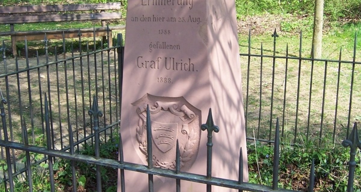 Ein Gedenkstein für Graf Ulrich, umgeben von einem Zaun, steht in einem Wald. Der Stein erinnert an den Tod von Graf Ulrich im Jahr 1388., © Natur.Nah. Schönbuch & Heckengäu Ein Gedenkstein für Graf Ulrich, umgeben von einem Zaun, steht in einem Wald. Der Stein erinnert an den Tod von Graf Ulrich im Jahr 1388., © Natur.Nah. Schönbuch & Heckengäu