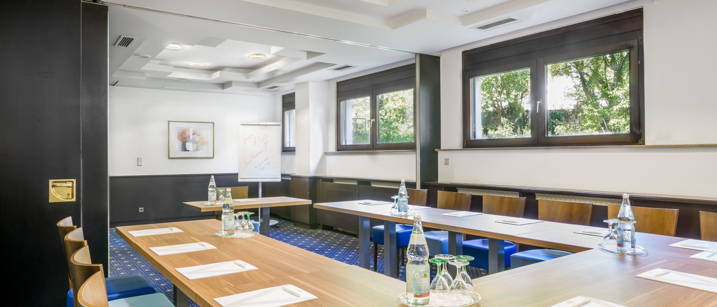 Tagungsraum mit U-förmiger Tischanordnung, Wasserflaschen und Schreibmaterial. Große Fenster und ein Flipchart im Hintergrund., © TOMAS