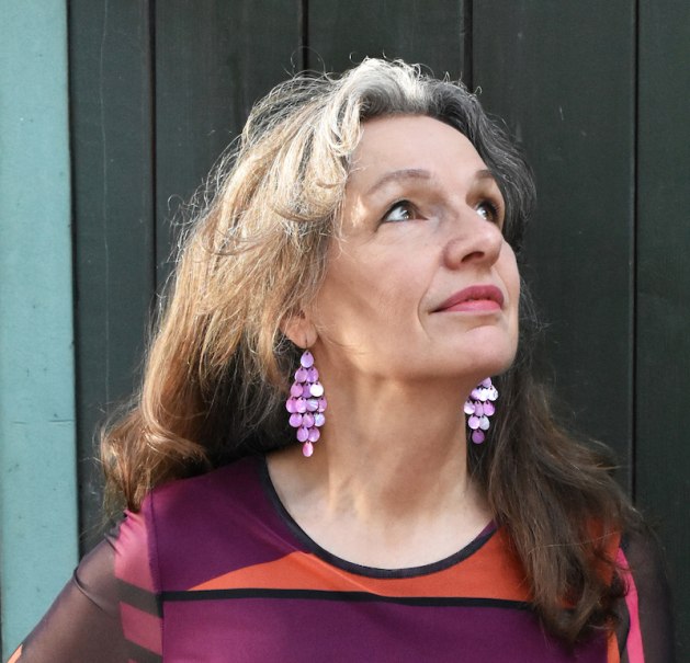 Eine Frau mit langen Haaren trägt auffällige lila Ohrringe und blickt nach oben. Sie trägt ein buntes Oberteil., © Dieselstrasse Kulturzentrum e.V. Eine Frau mit langen Haaren trägt auffällige lila Ohrringe und blickt nach oben. Sie trägt ein buntes Oberteil., © Dieselstrasse Kulturzentrum e.V.