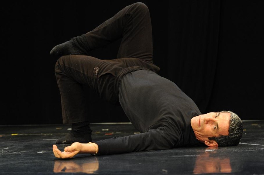 Ein Mann in schwarzer Kleidung f&uuml;hrt eine Feldenkrais-&Uuml;bung auf einem dunklen Boden aus. Er liegt auf dem R&uuml;cken mit einem angewinkelten Bein., &copy; &copy; Tanz- und Theaterwerkstatt
