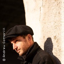 Eine Person mit Schiebermütze lehnt nachdenklich an einer hellen Wand, im Schatten eines Bogens. Foto von Andrew Cotterill., © links im Bild