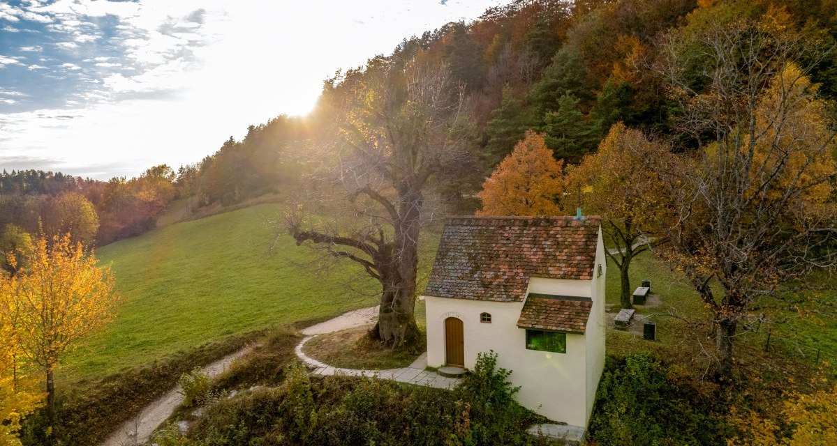 Die Reiterleskapelle steht in einer herbstlichen Landschaft, umgeben von bunten Bäumen und einer grünen Wiese. Die Sonne scheint durch die Wolken., © Foto Thomas Zehnder Die Reiterleskapelle steht in einer herbstlichen Landschaft, umgeben von bunten Bäumen und einer grünen Wiese. Die Sonne scheint durch die Wolken., © Foto Thomas Zehnder