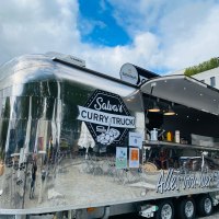 Ein glänzender Foodtruck mit der Aufschrift 'Salva's Curry Truck' steht im Freien vor einem modernen Gebäude., © Salva's Curry-Truck