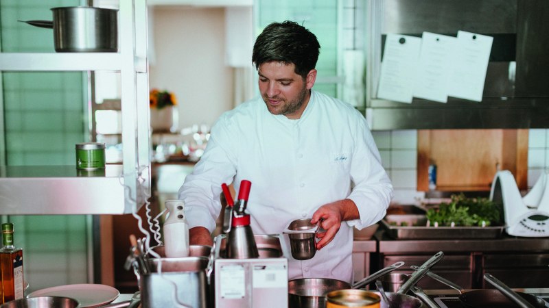 Ein Koch in weißer Uniform arbeitet konzentriert in einer professionellen Küche mit Töpfen und Utensilien., © Daniela Reske