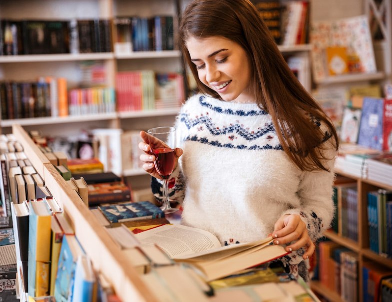 Frau in einer Buchhandlung liest ein Buch und hält ein Glas Rotwein. Sie trägt einen weißen Pullover mit Muster., © Osiander Vertriebsgesellschaft mbH & Co. KG Frau in einer Buchhandlung liest ein Buch und hält ein Glas Rotwein. Sie trägt einen weißen Pullover mit Muster., © Osiander Vertriebsgesellschaft mbH & Co. KG