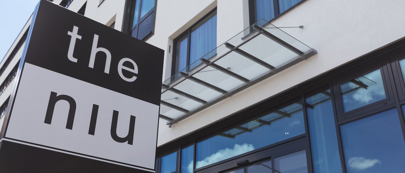 Moderne Gebäudeaußenfassade mit Glasvordach und dem Schild 'the niu' im Vordergrund., © the niu Form - Novum Hospitality
