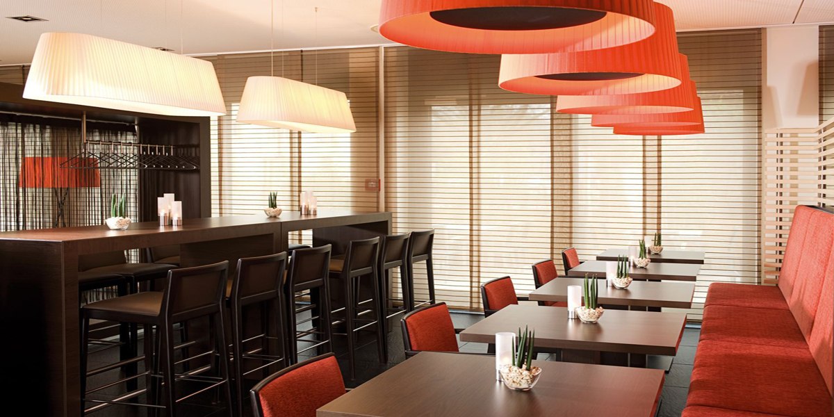 Moderner Frühstücksraum mit roten Lampen, Holztischen und Stühlen. Große Fenster mit Jalousien sorgen für viel Licht., © NH Ludwigsburg