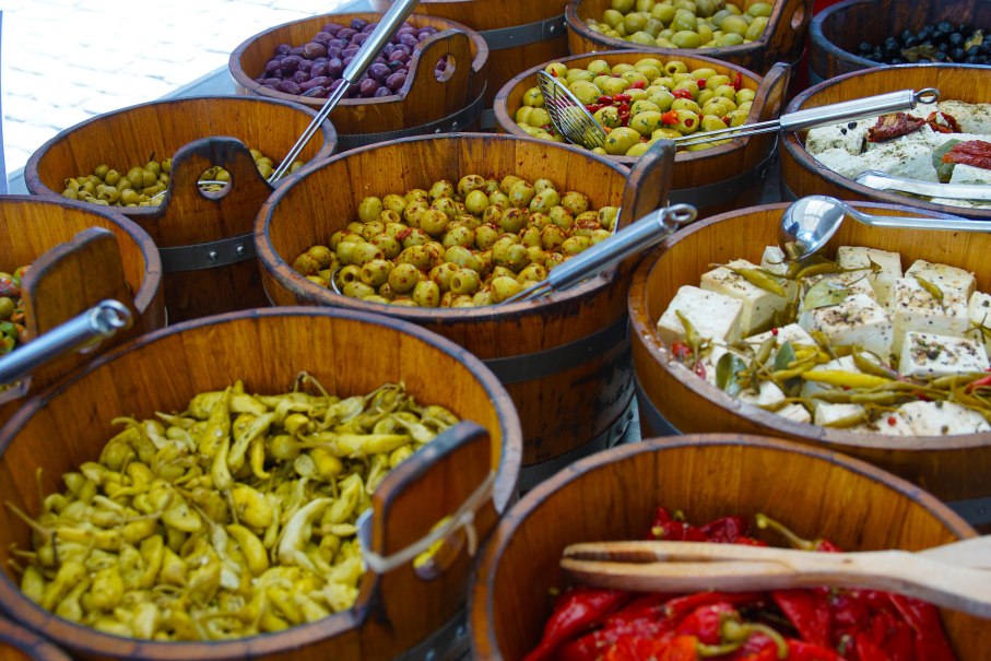 Holzfässer mit verschiedenen Antipasti wie Oliven, Peperoni und Käse auf einem Marktstand. Die Vielfalt der Farben und Texturen ist sichtbar., © T. Krytzner