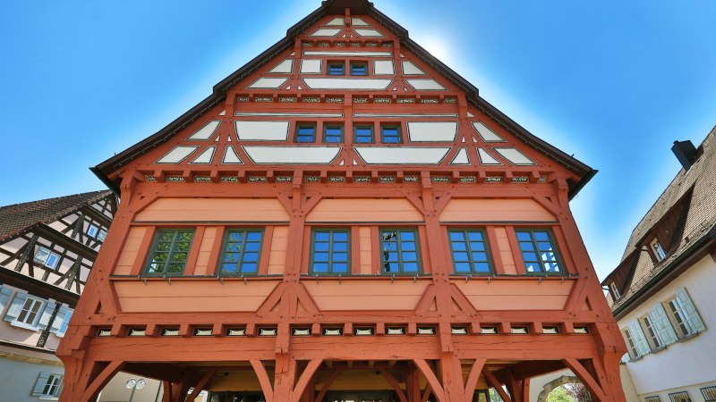 Fachwerkhaus mit rotem Holzrahmen und blauen Fenstern, umgeben von weiteren Fachwerkgebäuden, unter klarem, blauem Himmel., © SMG Achim Mende