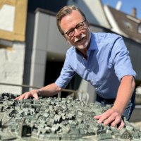 Ein Mann in einem blauen Hemd beugt sich lächelnd über ein detailliertes Stadtmodell. Im Hintergrund sind Gebäude und blauer Himmel zu sehen., © WTM GmbH Waiblingen