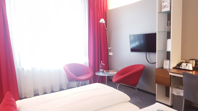 Hotelzimmer mit Doppelbett, roten Sesseln, kleinem Tisch, Wand-TV und Schreibtisch mit Kaffeemaschine. Helle Vorh&auml;nge und rote Akzente., &copy; Vienna House Easy Mo. Stuttgart