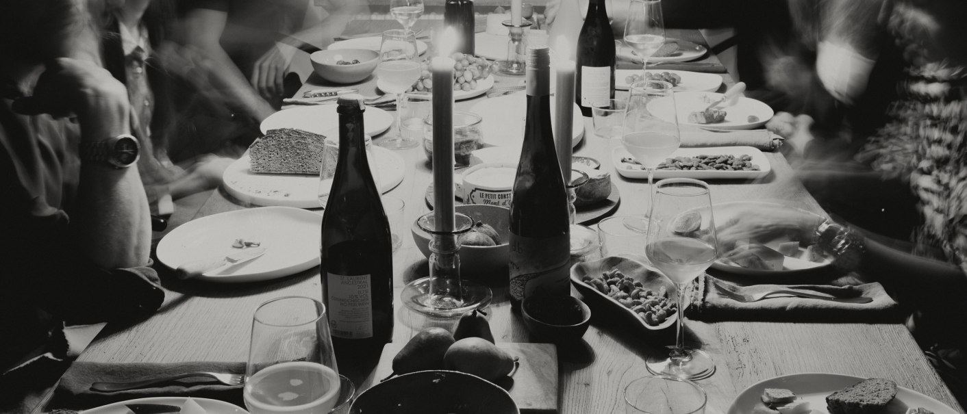 Ein Esstisch mit Kerzen, Weinflaschen und Tellern. Menschen sitzen um den Tisch, die Szene ist in Schwarz-Weiß gehalten., © Peter Oliver Wolff / DAS EDITH